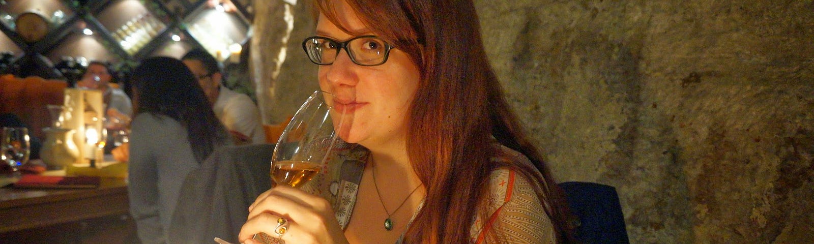 Author with long red hair and glasses drinking wine in a basement wine cave in Budapest during her travels