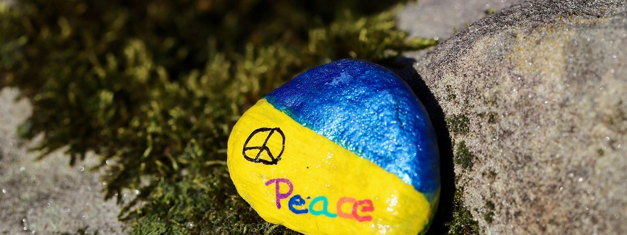 A small rock painted in the colors of the Ukrainian flag (blue and yellow) with a peace sign and the word “peace” written in colorful letters on the yellow part.