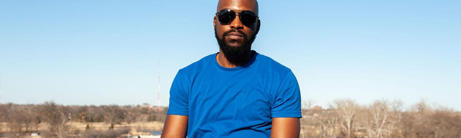 Erik is a Black man is sitting on the ledge in the sun, with the Forest Park Art Hill fountains in the background. It’s a bright sunny day and he is bald, wearing sunglasses, has a goatee, and is wearing a blue t-shirt with blue jeans. He has a neutral expression on his face.