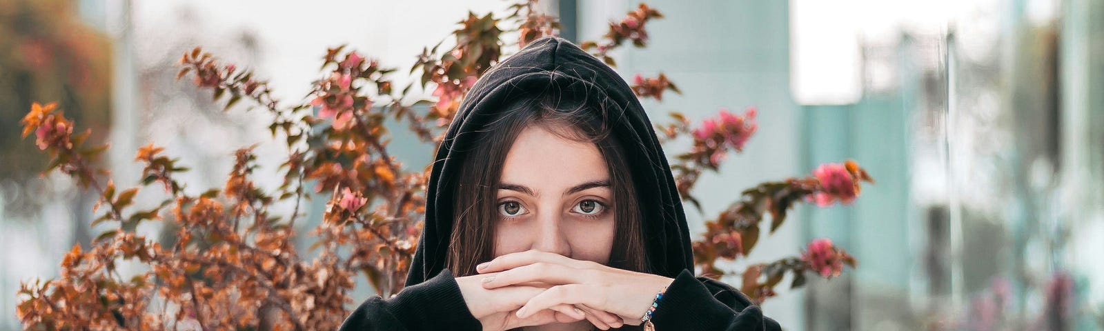 Sadly reflective young woman in a dark hoodie sitting with her elbows on the table, hands clasped in front of her mouth.