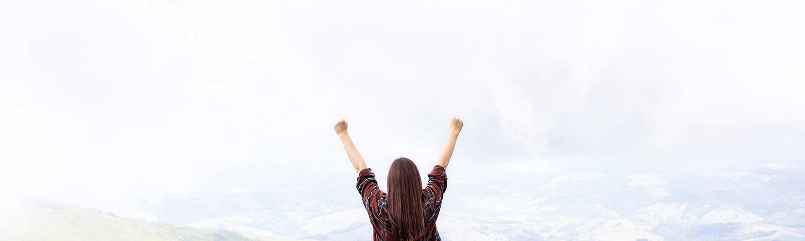 Back profile of a woman standing in front of mountain landscape. Both hands are in the air as if she’s letting things go.