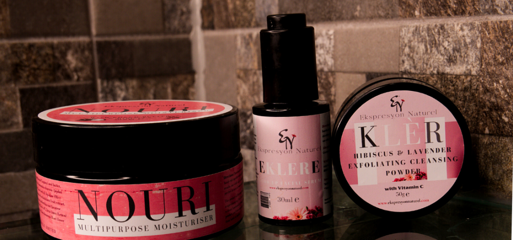 Three items on a reflective table. One round pot with the word Nouri on it written in black text with a pink sticker on the front and on the lid. One dark coloured bottle with pink label and black and white lettering. Another round pot with pink black and white lettering.