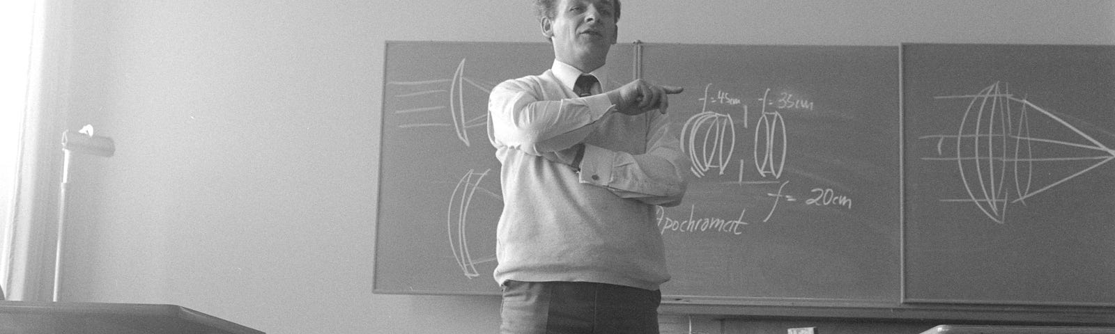 A teacher pointing at the class, whilst standing in front of blackboards.