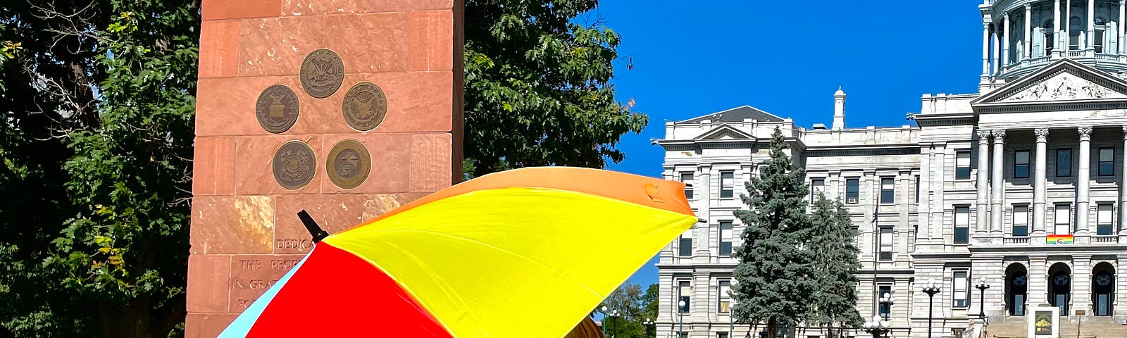 The author is wearing rainbow angel wings and is carrying a rainbow parasol with the words “Get Your Gay On!” written on it. He is standing in front of the Denver Capital Building.