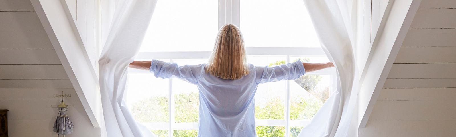 A woman is throwing the white curtains wide on her window, back to us