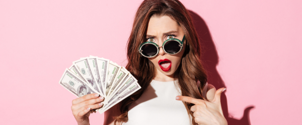 A woman with cash and a surprised look on her face. She is pointing to the cash and looking over the brim of her eye glasses.