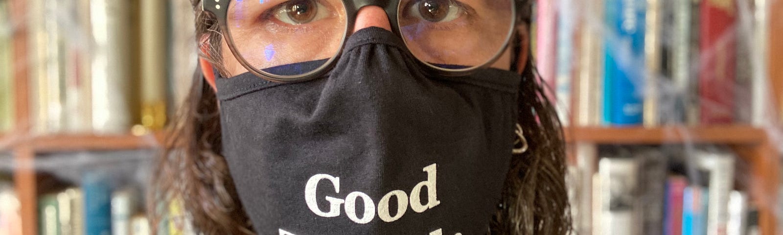 Close-up of me, wearing glasses, in front our bookshelf, my face half covered by a mask reading, “Good Trouble”.