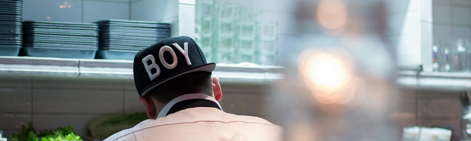 A chef wearing a cap with the word ‘boy’ in large white print has his back to the camera as he works in a restaurant kitchen, probably preparing food.