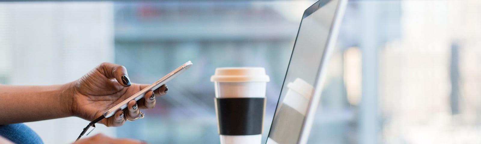 Photo of a woman typing on her laptop, while holding a phone in her left hand, with glasses and coffee cup on the table too.