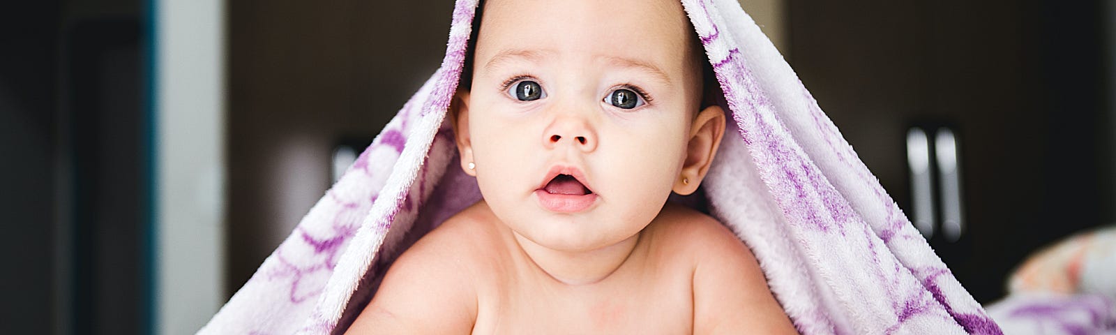 Baby on belly pushing up with their arms almost smiling with a purple and white blanket draped over the sides of their head.