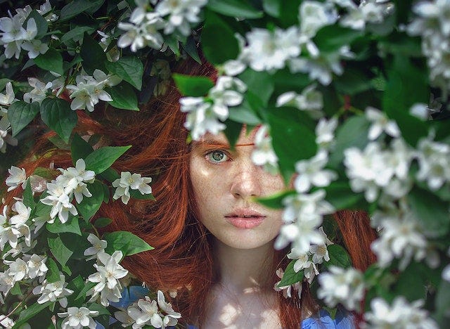 A red-headed woman partially obscured by green leaves and white flowers.