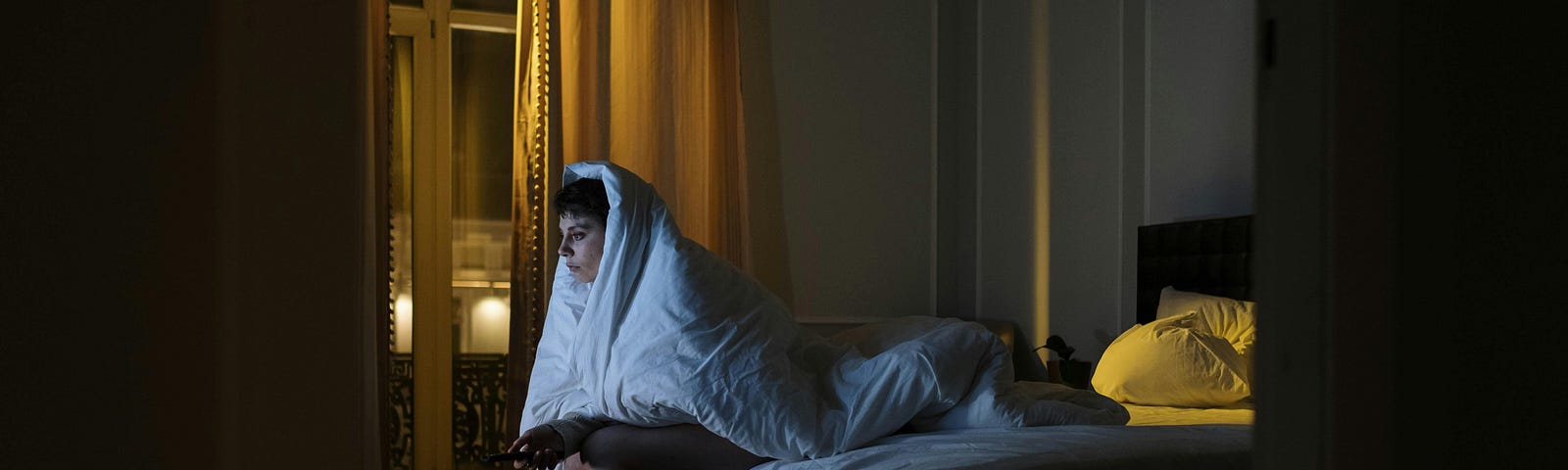photo-of-a-woman-sitting-on-the-bed-while-covered-by-a-white-blanket