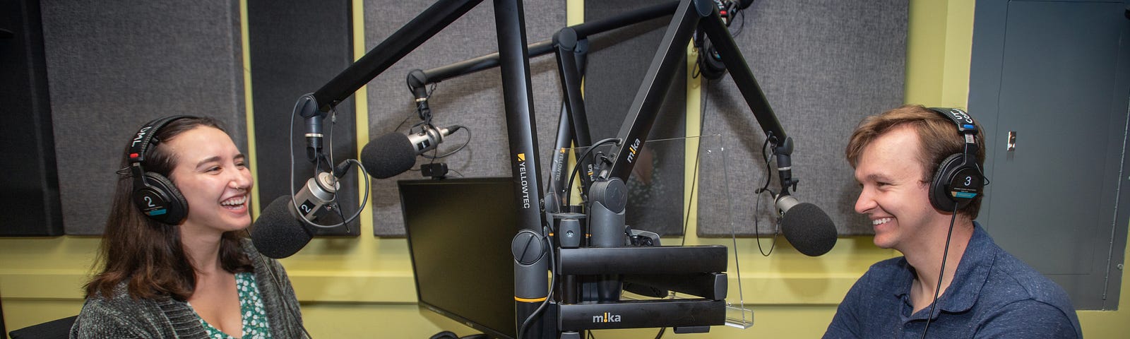 Two people sit in a recording booth talking into microphones.