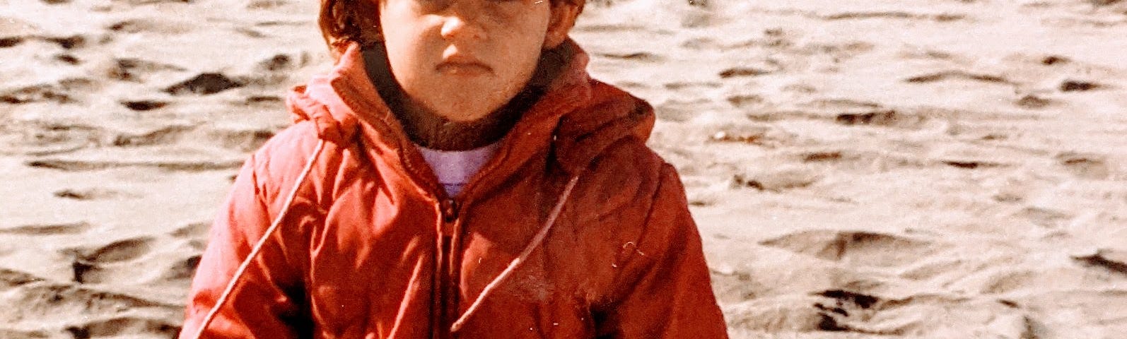 An old picture of a little girl with white skin, blonde hair, pink jacket, holding a bucket and shovel at the beach.