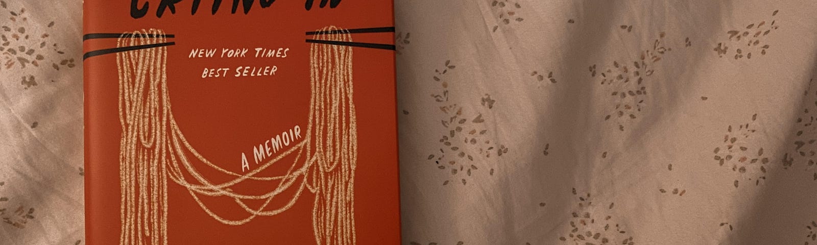 The book “Crying in H Mart” by Michelle Zauner is laying flat on a white sheet with a subtle floral pattern .