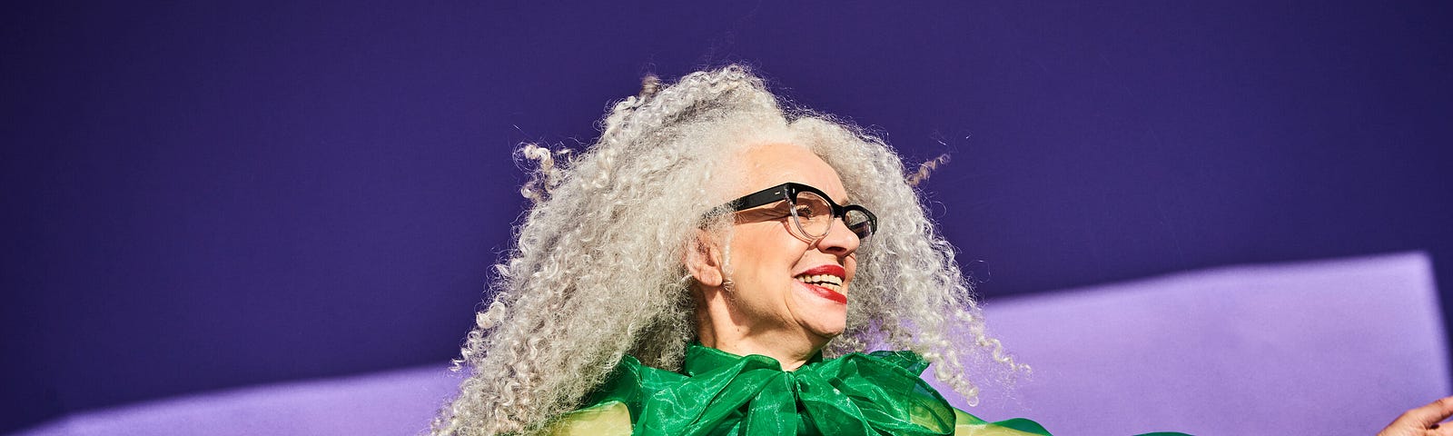 Portrait of a woman with gray curly hair and glasses wearing a green shirt against a purple background