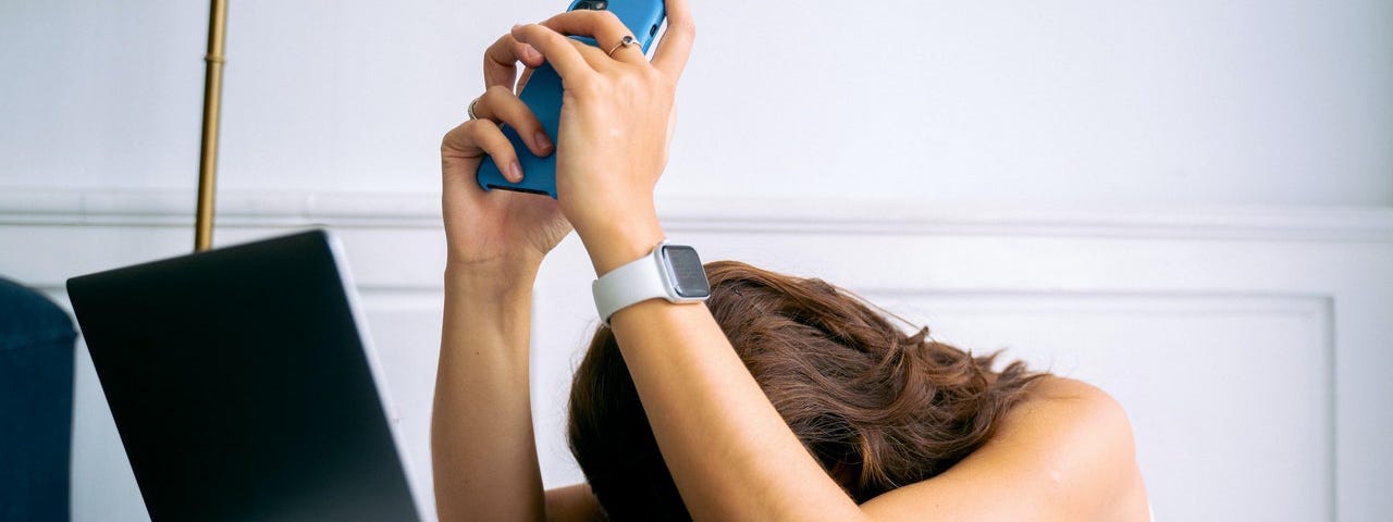 A woman with her head face down on a white table while holding her phone and a her laptop in front of her