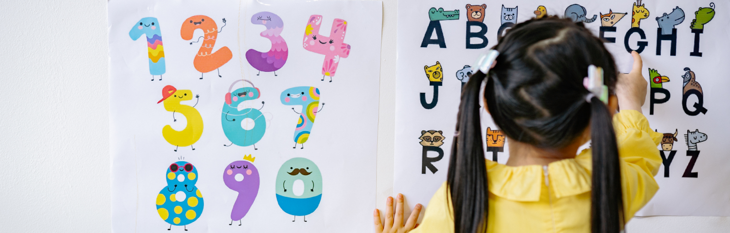 Image shows two charts, one containing numbers and the other containing letters. A preschool girl points at the chart containing letters, her back facing the viewer.