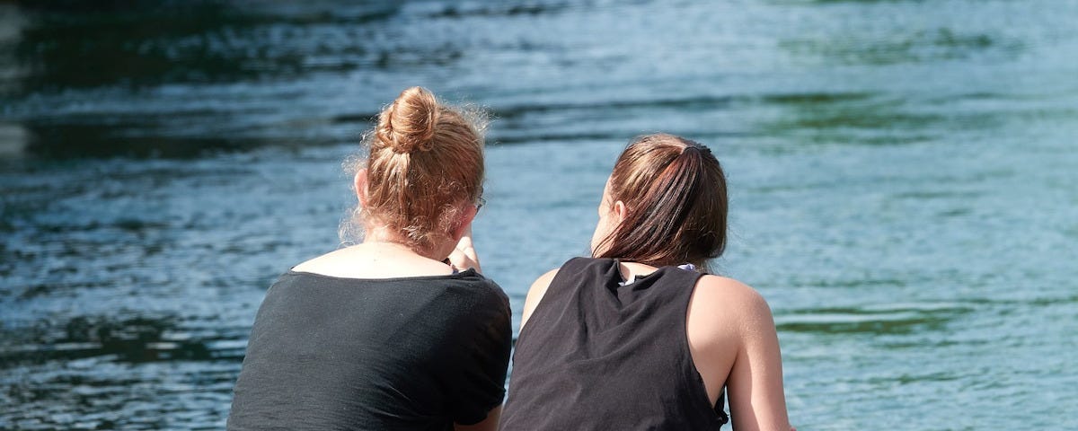 A rear view of two women wearing black tops sitting on a bench. One is looking out over a lake, the other is looking at her.