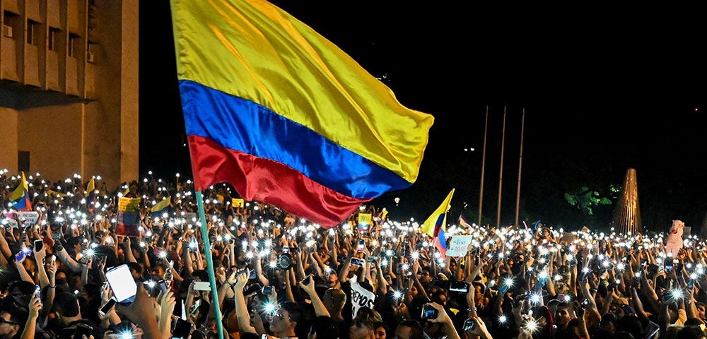 People protesting at the streets, holding their cellphones up. One big Colombian flag in the front.
