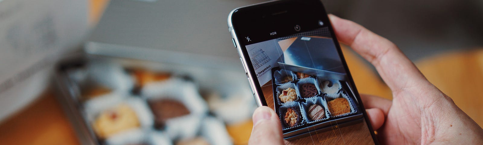 A pair of hands holding a smartphone take a picture of baked goods in a box. The image can be seen on the smartphone display.