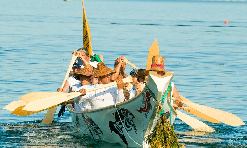 A light colored traditional canoe with Native American artwork on the side is shown. There is a bundle of greenery hanging on the front of the canoe and a yellow flag with lettering is attached to the back. Nine people are in the boat using traditional paddles to move the boat through the water. Several of the people in the boat wear traditional woven hats.