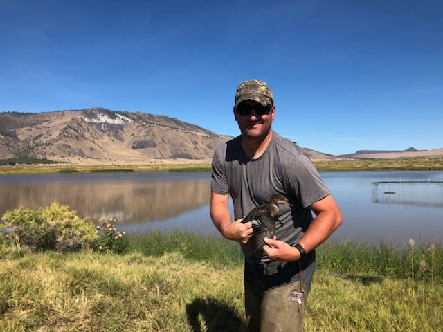 Joe poses, wearing a ball cap with a backpack. a dry brown landscape stretches back behind him. The sky above is a vivid blue.