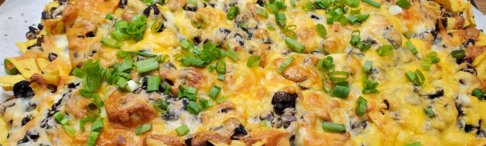 A big pile of tortilla chips, taco meat, sliced mushrooms and cheese topped with green onions on a parchment paper line baking sheet.