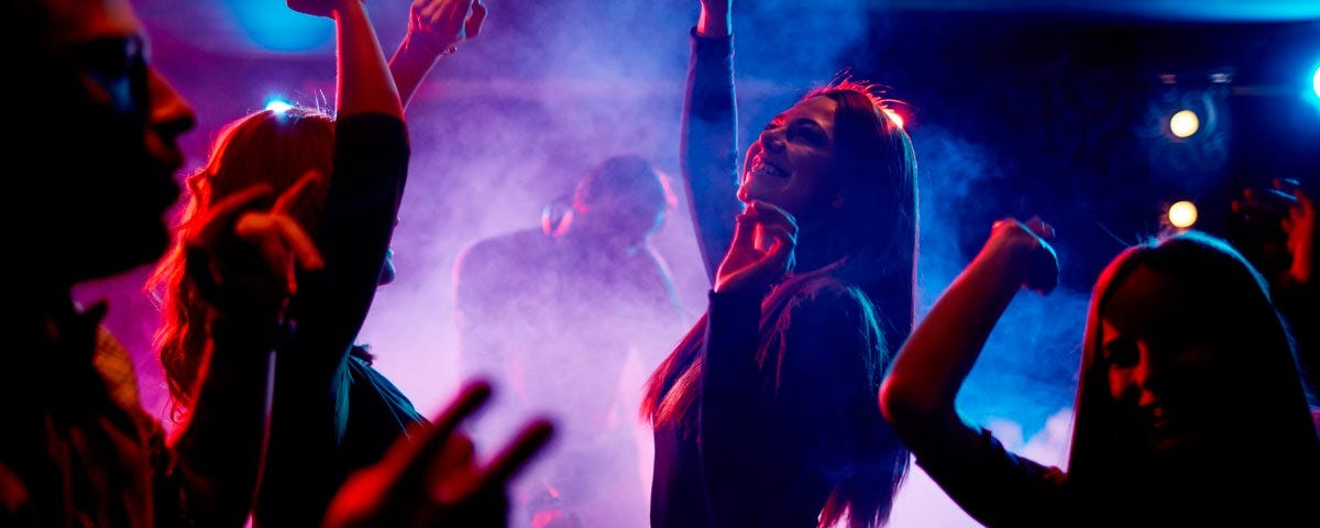 Silhueta de várias pessoas dançando com um ambiente escuro e o fundo iluminado com luzes azuis e roxas e uma fumaça.