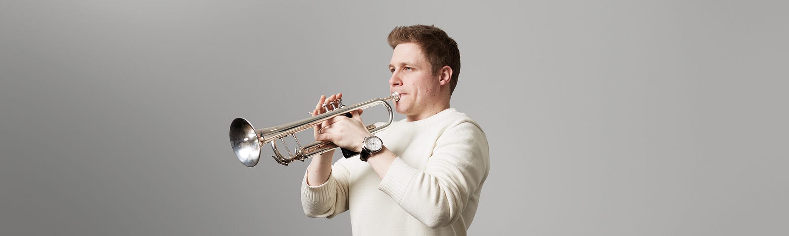 Philip Cobb playing trumpet wearing a white jumper, in front of a plain grey background