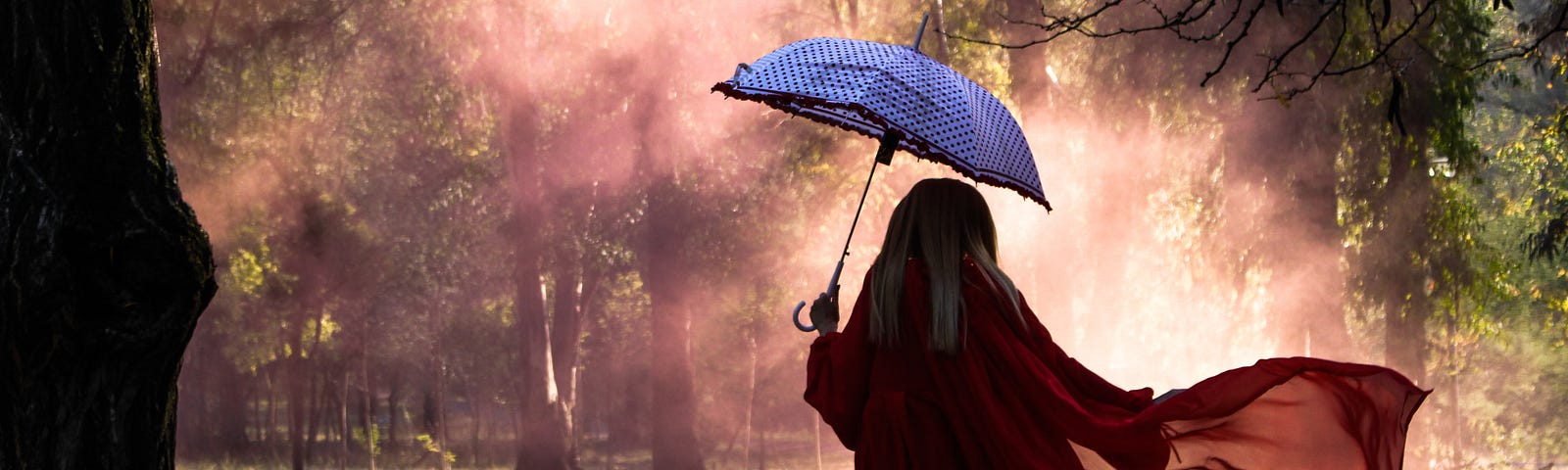 A woman wearing red, carrying an open blue umbrella approaching a patch of sunlight in the park.
