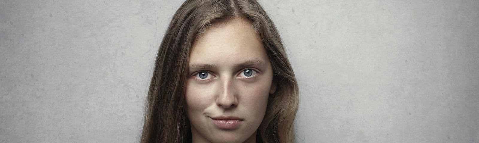 young white woman with blond hair smirking. She’s wearing a grey tank top against a grey backdrop