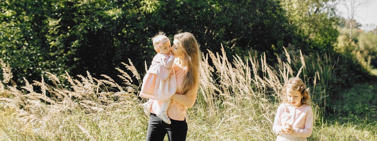 A young mother holds her baby and kisses her as the older daughter stands off to the side, lost in thought. They are outside in a field.