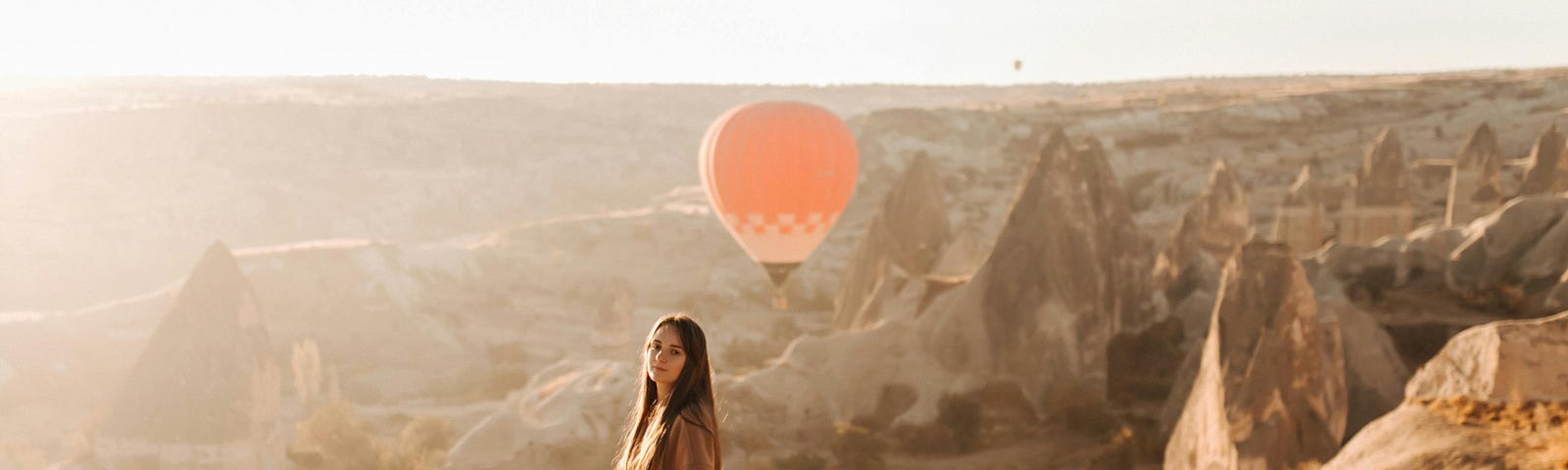 woman wearing brown coat standing outside, on a cliff. orange hot hair balloon in the sky