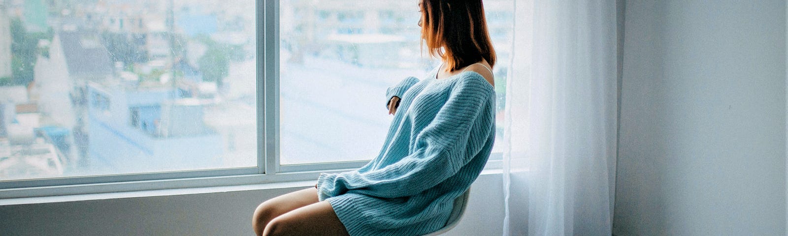 somber girl with brown hair wearing long sleeve blue sweater sitting on chair in empty room looking out the window