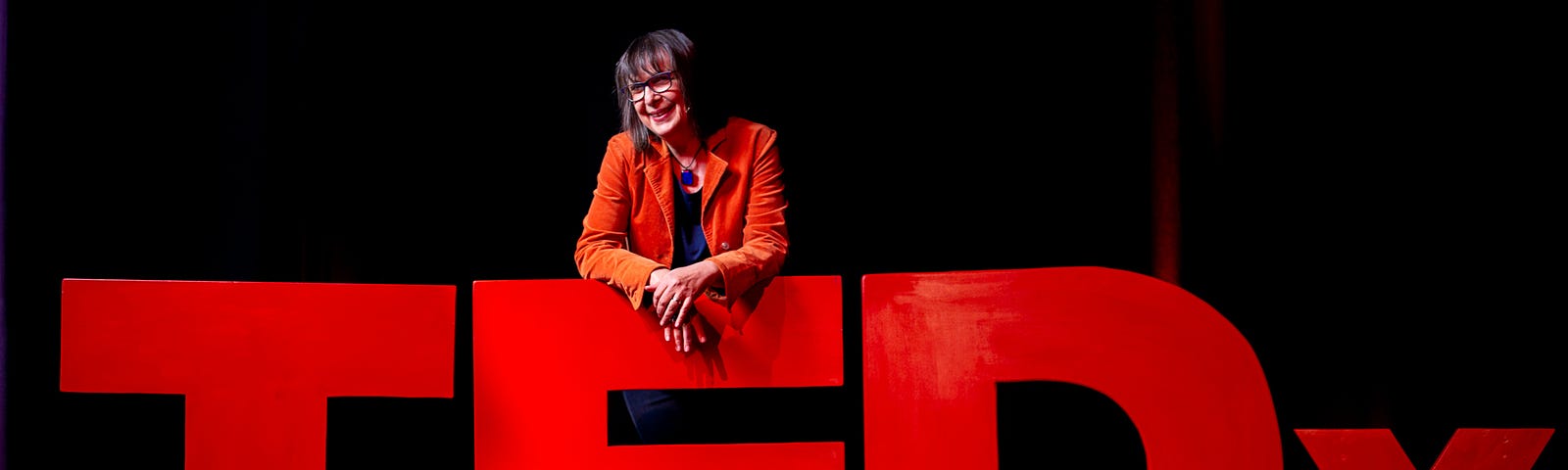 The author Trisha Lewis on the TEDx stage. Leaning over the ‘E’ of the giant red TEDx blocks.