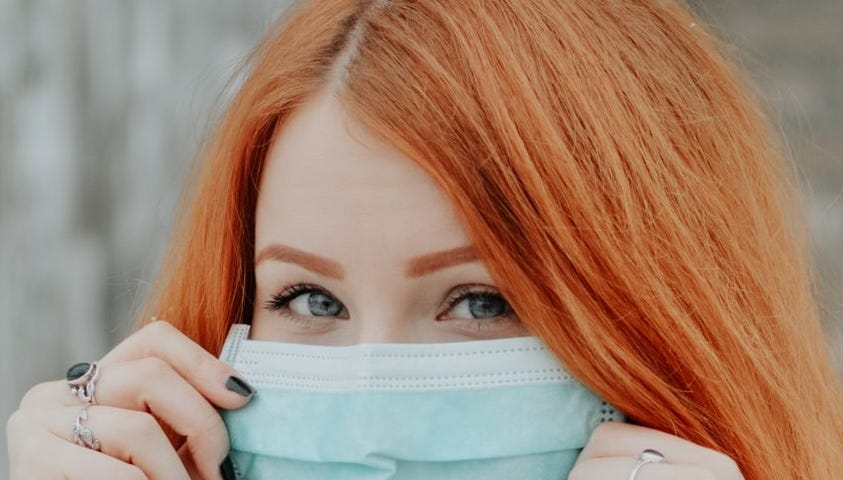 Young woman with red hair, holding a mask up to her face.