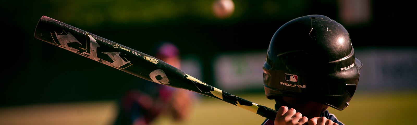 Young boy in baseball uniform and helmet holds a baseball bat ready to swing as a baseball comes flying at him