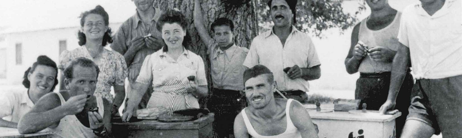 Seven men and three women group together in front of a wooden table and two ice cream carts. A man in the center of the image is smiling and wearing a white tank top and shorts. To his left, another man in a white tank top and shorts takes a bite of ice cream.