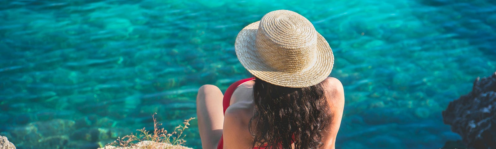 A woman with her back to the camera sits on some rocks facing the ocean. She is wearing a red bikini and has long, curly brown hair tucked into a straw hat.