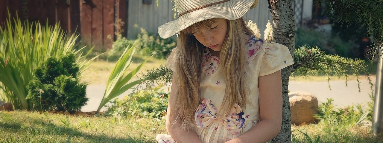 Young girl with blond hair and hat reading a book while sitting on grass and resting on a tree. A second book with an apple on it is beside her.