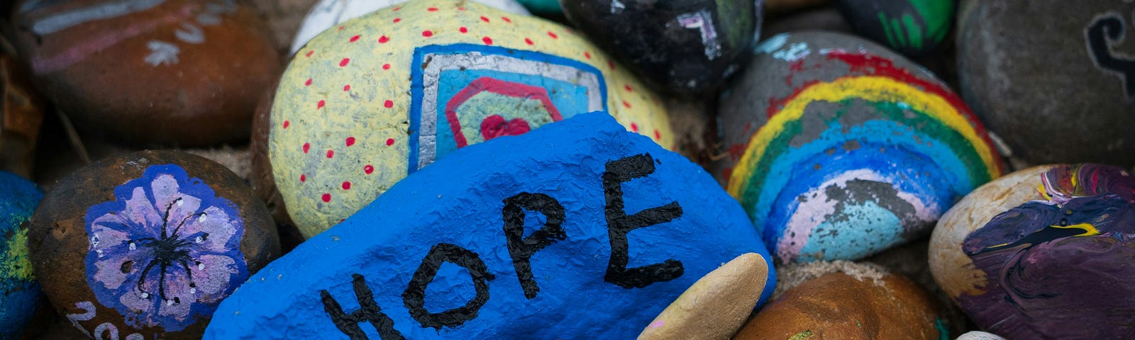 A variety of painted rocks with the focus being on a blue rock in the middle with black letters spelling HOPE.