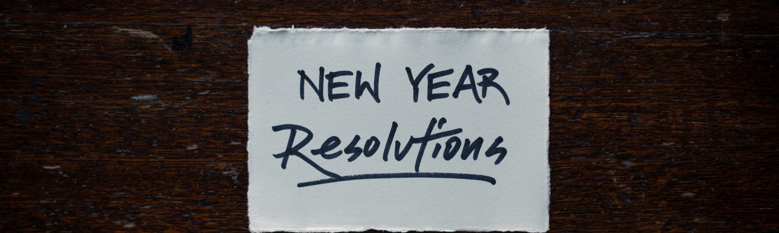 An index card with New Year Resolutions written on it, where Resolutions is underlined. Under index card is a capped pen with a white-and-gold marble design.