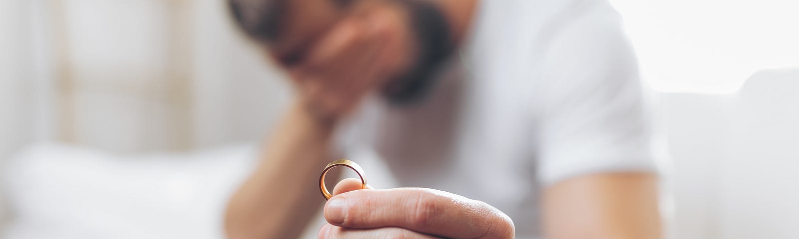 A man holds his head in grief. In front of him, a marriage band is held outstretched in his hand.