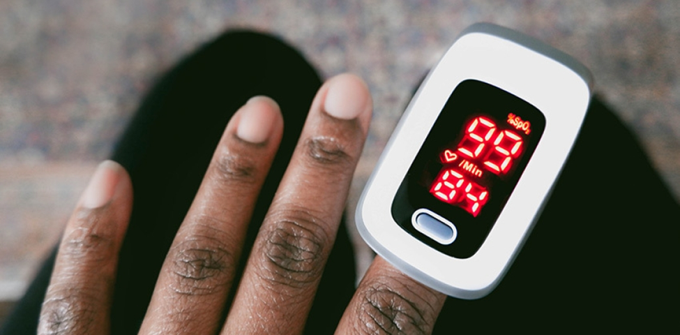A Black person’s hand with a pulse oximeter on their finger, reading an oxygen saturation of 99% and 84 beats per minute