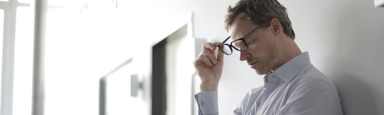 Man in dress shirt leans up against wall with eyes closed and removes eyeglasses.