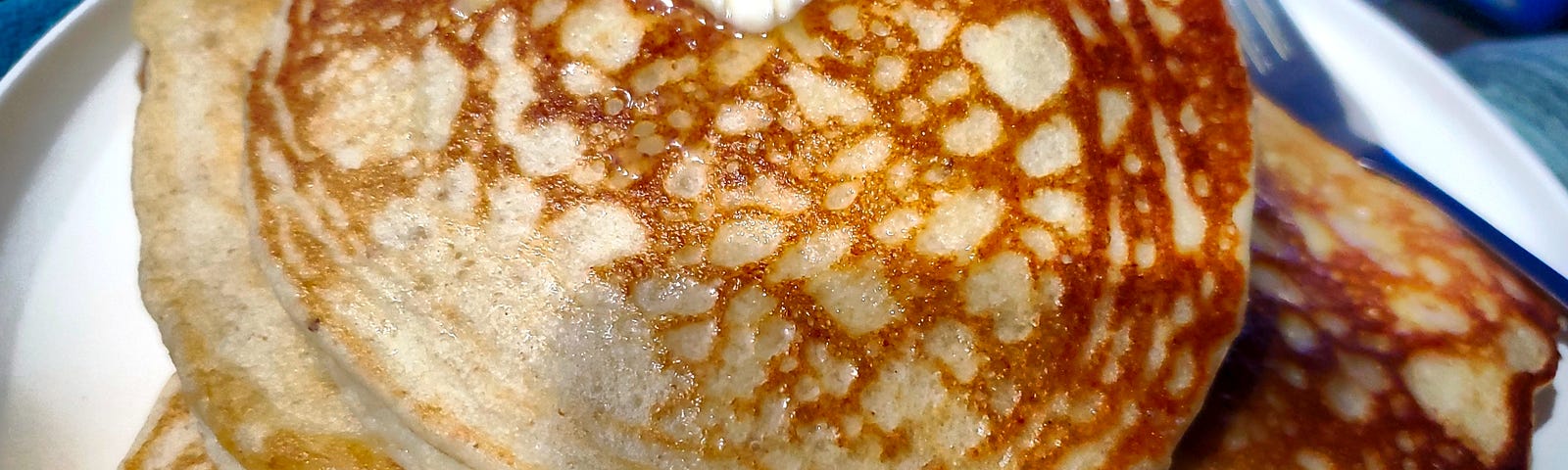 Looking down onto a white plate with large pancakes stacked on it. The top pancake has a pat of butter melting on it.