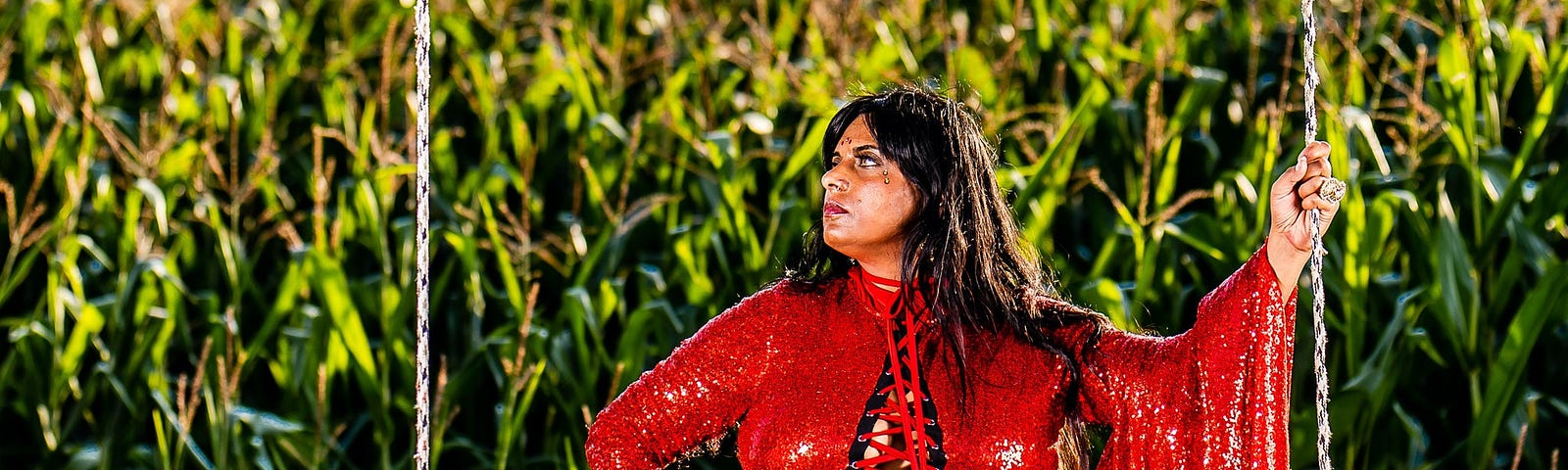 Fierce-looking BBW POC in a draped red dress standing before a cornfield. They have a foot in red high-heeled shoe planted staunchly upon an old tree swing.