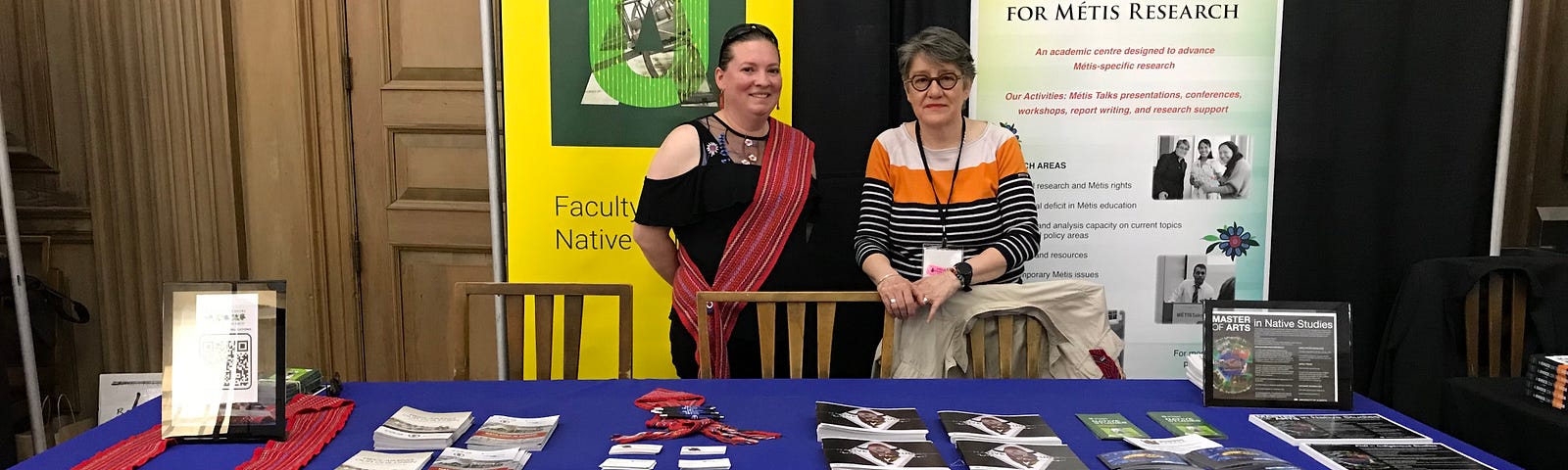 2 women, Amanda Evans and Nathalie Kermoal, stand behind a table that reads “Rupertsland Centre for Metis Research” and in front of banners for the Faculty of Native Studies and RCMR.