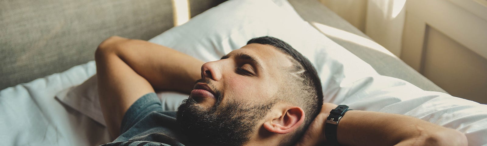 man lying on a comfortable bed with arms folded behind his head, taking a daytime nap and grinning